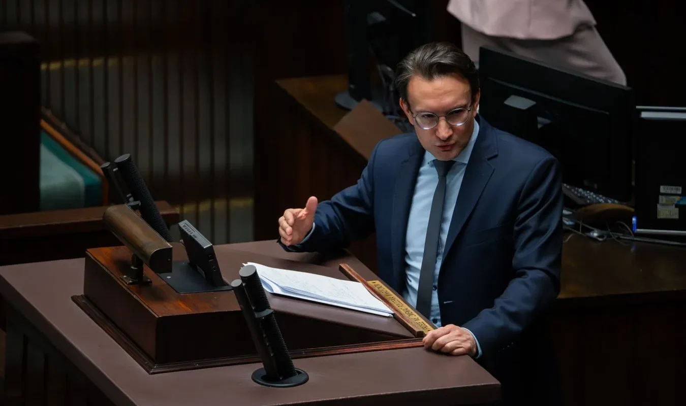 Mężczyzna w granatowym garniturze przemawia z notatkami przy pulpicie w sali plenarnej.