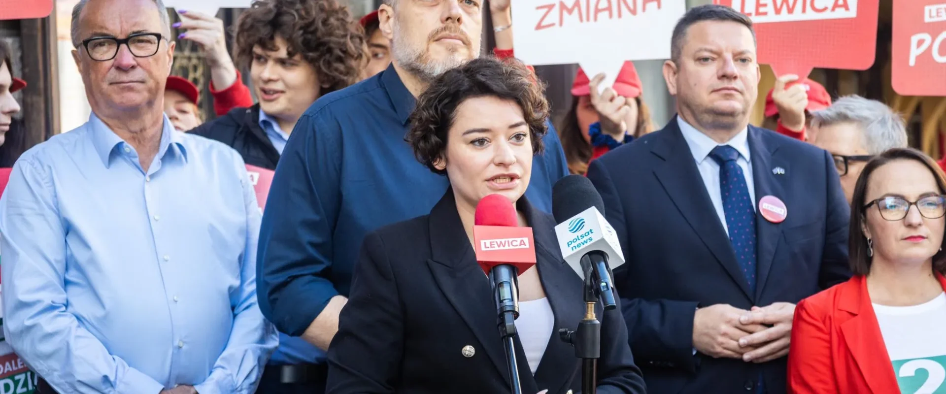 Polityczka Lewicy przemawia na konferencji prasowej, stojąc przy mikrofonach z logotypami Lewicy i Polsat News, w tle tłum z plakatami wyborczymi.