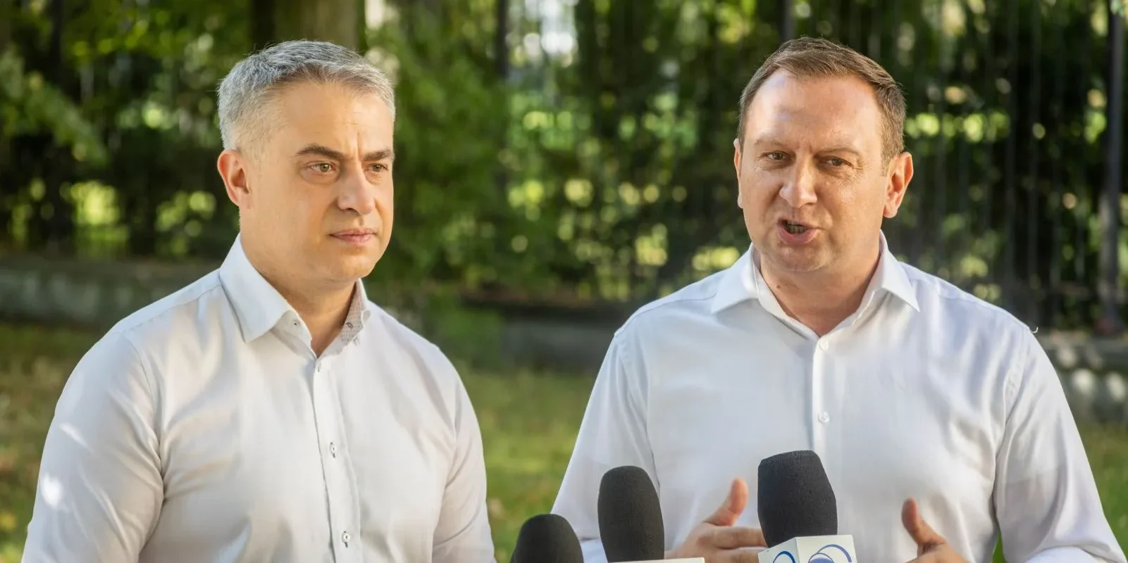 Dwóch mężczyzn w białych koszulach przemawia na konferencji prasowej na świeżym powietrzu, stojąc przy mikrofonach z logotypami TVP Info i TVN24, w tle zieleni, w kontekście referendum.