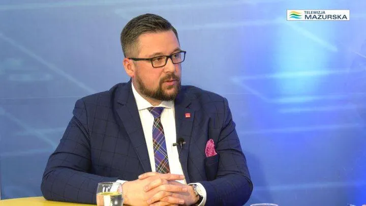 Mężczyzna w granatowej marynarce, białej koszuli i kraciastym krawacie siedzi przy stole w studiu telewizyjnym, z mikrofonem przy koszuli i różową poszetką w klapie, na tle niebieskiego ekranu z logiem Telewizja Mazurska.