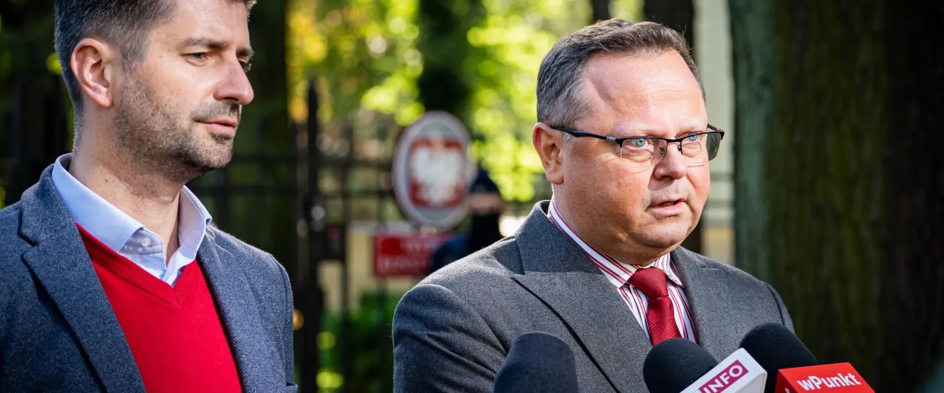 Dwóch mężczyzn w garniturach na konferencji prasowej dotyczącej odwołania ambasadorów; po prawej mężczyzna z okularami odpowiada na pytania, stojąc przy mikrofonach z logotypami TVN24, TVP Info i WPunkt, w tle zielone drzewa.