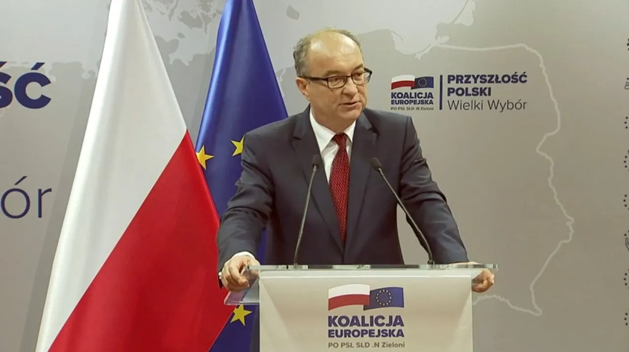 Mężczyzna w garniturze przemawia z podiumem na tle flag Polski i Unii Europejskiej oraz logo Koalicji Europejskiej.