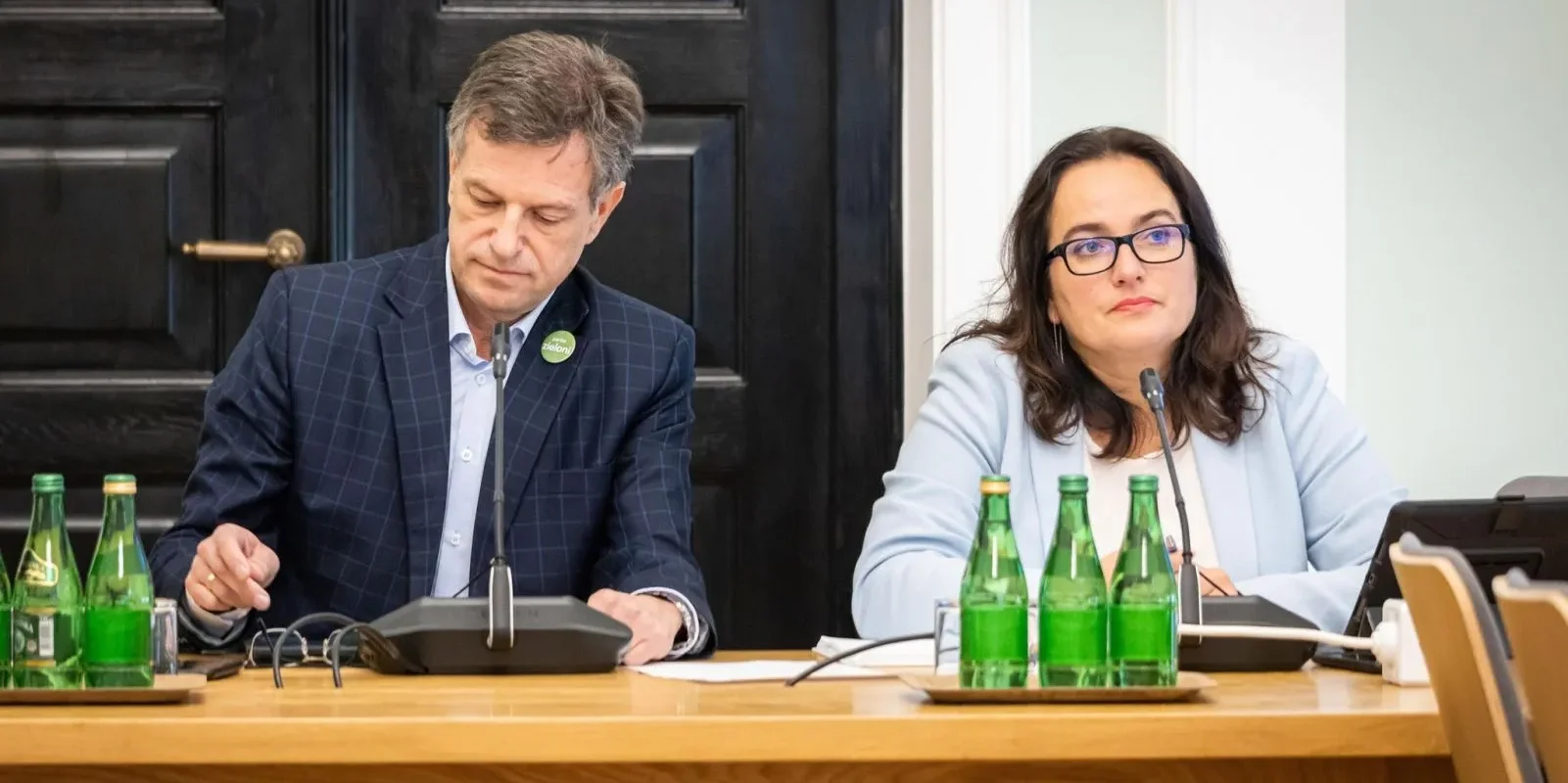 Na zdjęciu widać dwóch uczestników spotkania przy stole, z butelkami wody na blacie oraz telefonami konferencyjnymi, w skoncentrowanej atmosferze.