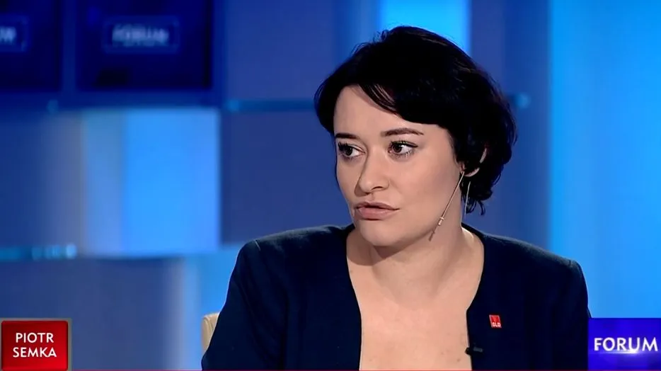 Kobieta w czarnym garniturze siedzi w studiu telewizyjnym, z mikrofonem na klipsie, na tle niebieskiego oświetlenia i ekranów.
