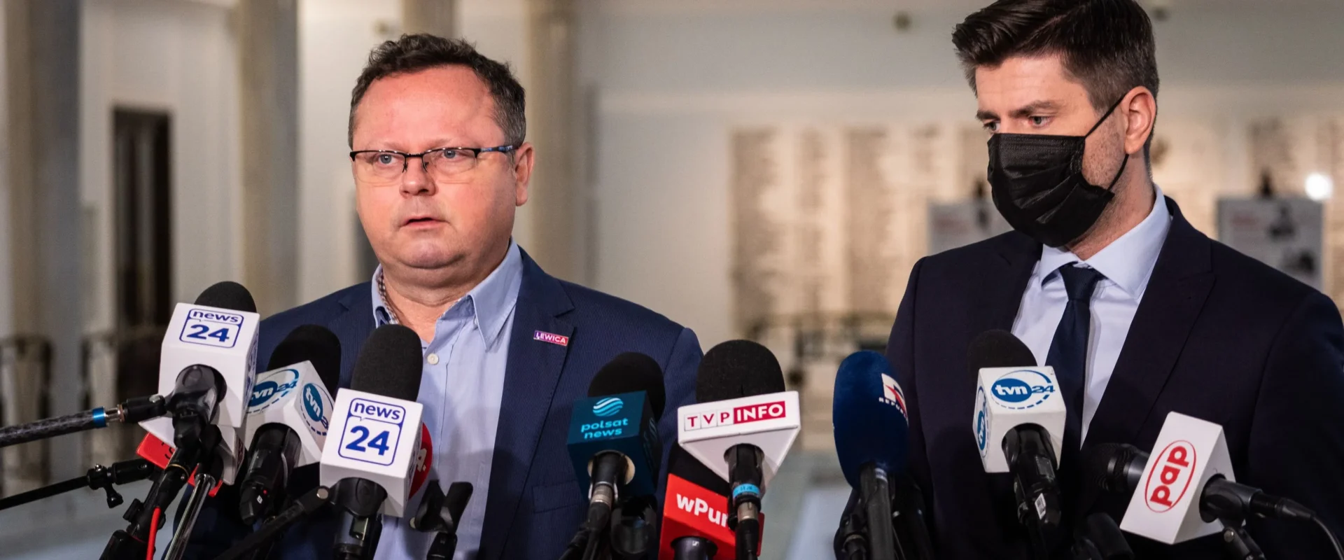 Dwóch mężczyzn w garniturach podczas konferencji prasowej, otoczonych licznymi mikrofonami z logotypami mediów; jeden bez maseczki, drugi w czarnej maseczce.
