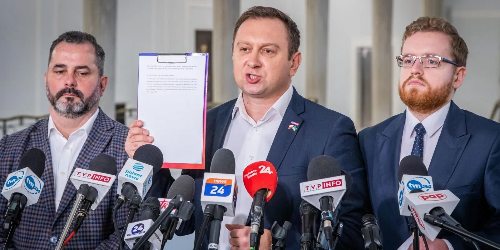 Na zdjęciu trzech mężczyzn stoi przed mikrofonami podczas konferencji prasowej, a jeden z nich prezentuje dokument.
