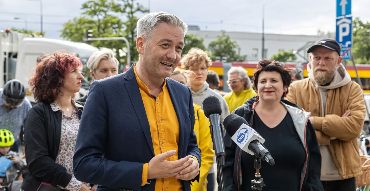 Mężczyzna w niebieskiej marynarce i pomarańczowej koszuli przemawia do zgromadzonego tłumu, któremu towarzyszą różnorodne postacie, w tym kobieta w czarnej bluzie.