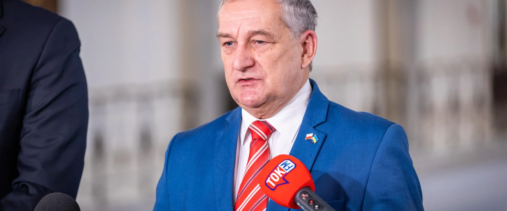 Mężczyzna w niebieskim garniturze i czerwonym krawacie przemawia do reporterów podczas konferencji prasowej, stojąc obok mikrofonów.