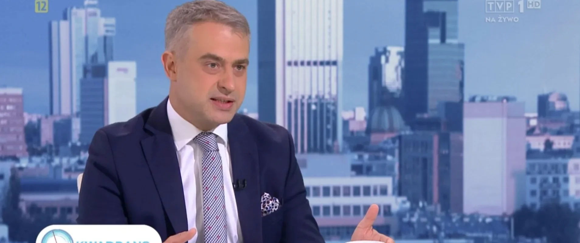 Mężczyzna w garniturze prowadzi program w studiu telewizyjnym, z widokiem na miasto w tle, a na stole stoi filiżanka i widoczny jest logotyp Kwadrans Polityczny.