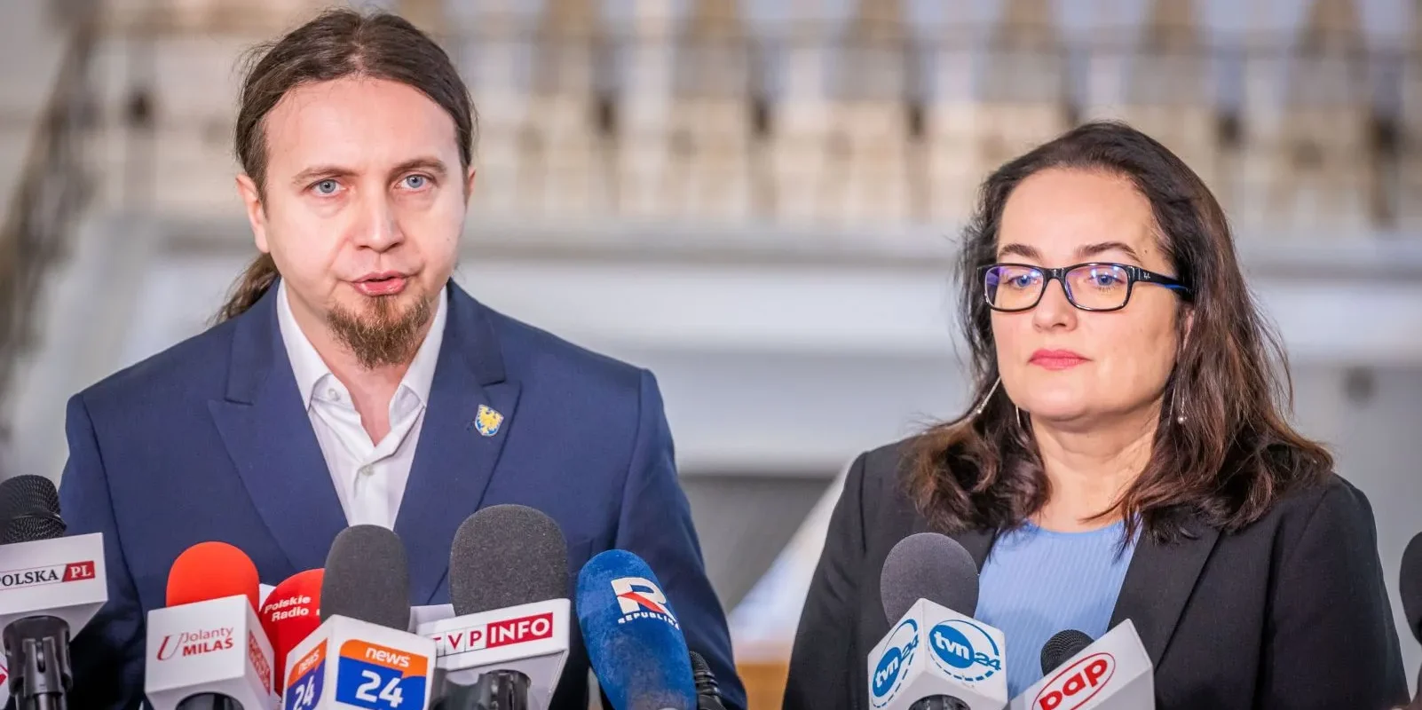 Na zdjęciu znajdują się dwie osoby stojące przed mikrofonami, podczas konferencji prasowej w formalnym otoczeniu.