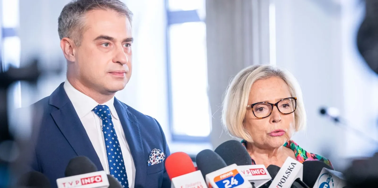 Na pierwszym planie dwie osoby — mężczyzna w granatowym garniturze i kobieta w okularach — stoją przy licznych mikrofonach podczas konferencji prasowej.