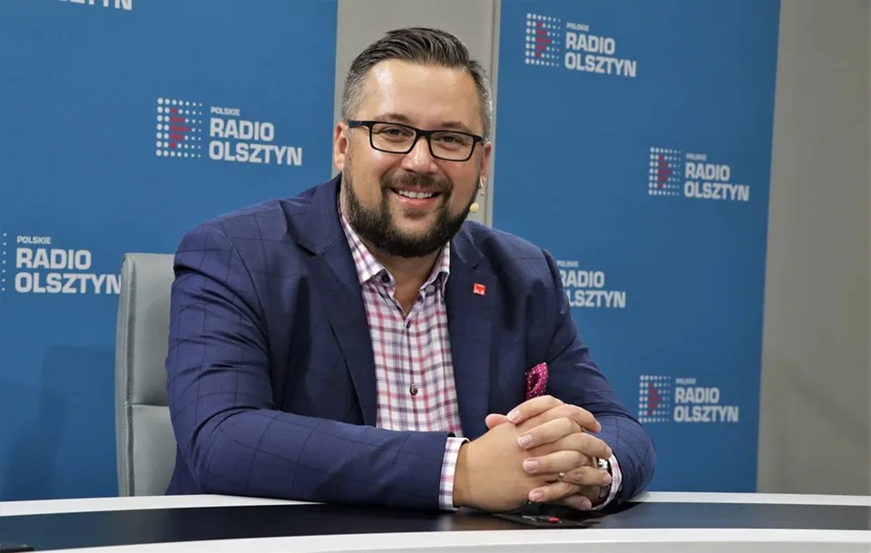 Mężczyzna w granatowym garniturze siedzi przy biurku w studiu radiowym, na tle niebieskiego banneru z logo Radio Olsztyn.