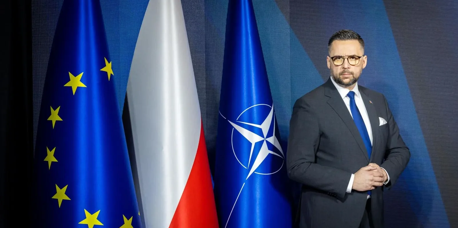 Mężczyzna w garniturze stoi przed flagami Unii Europejskiej, Polski i NATO.