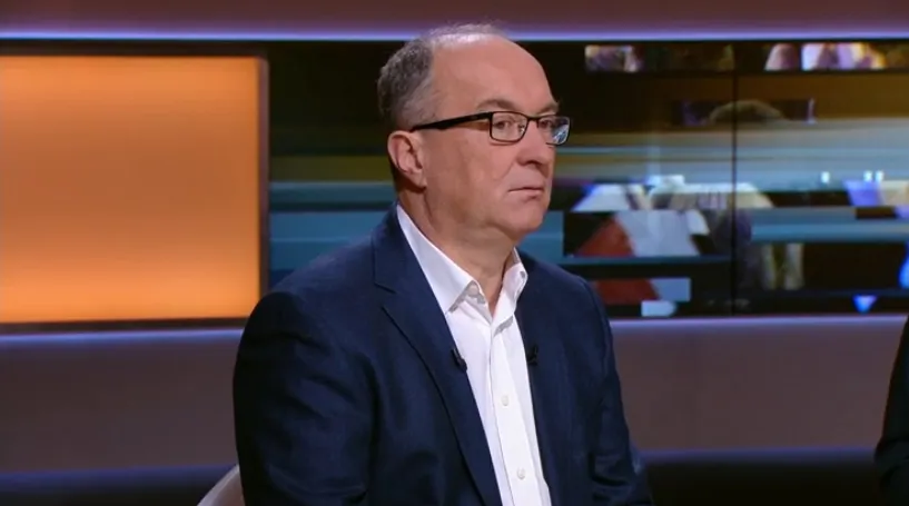 Mężczyzna w granatowym garniturze i okularach siedzi w studiu telewizyjnym, na tle paneli i ekranów.