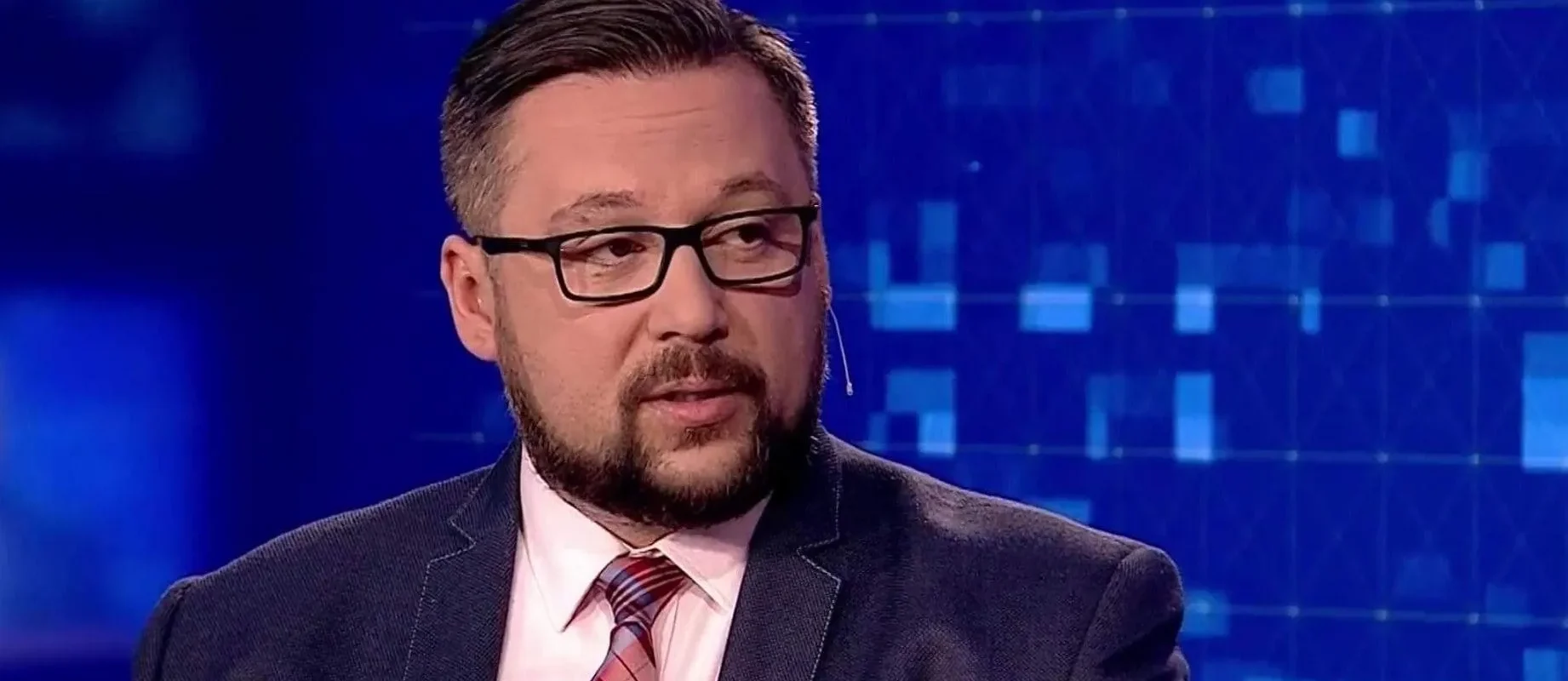 Mężczyzna w garniturze i okularach, z brodą i mikrofonem na uchu, siedzi w studiu telewizyjnym z niebieskim, cyfrowym tłem.
