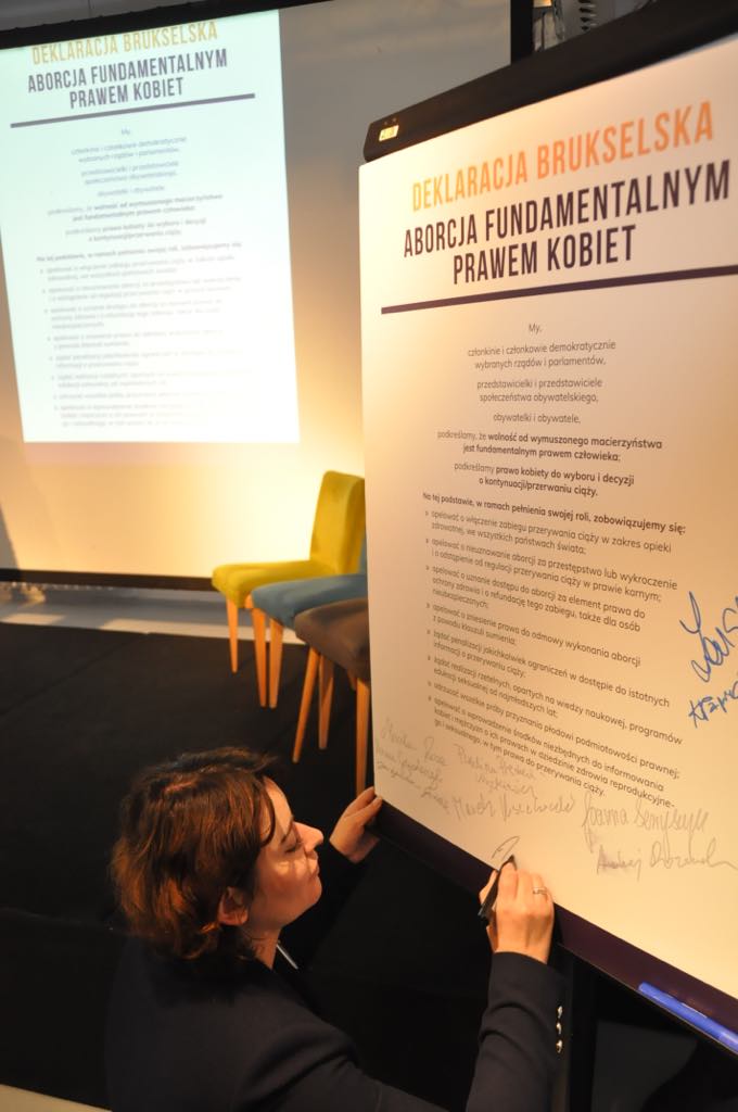 Kobieta podpisuje duży plakat z tytułem „Deklaracja Brukselska – Aborcja fundamentalnym prawem kobiet” podczas prezentacji na scenie.