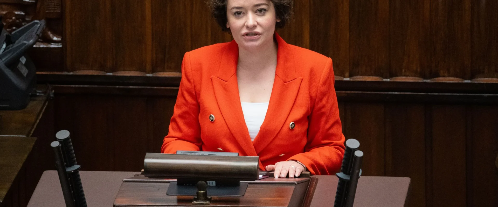 Kobieta z krótkimi, kręconymi włosami w czerwonej marynarce siedzi przy mównicy w sali plenarnej.