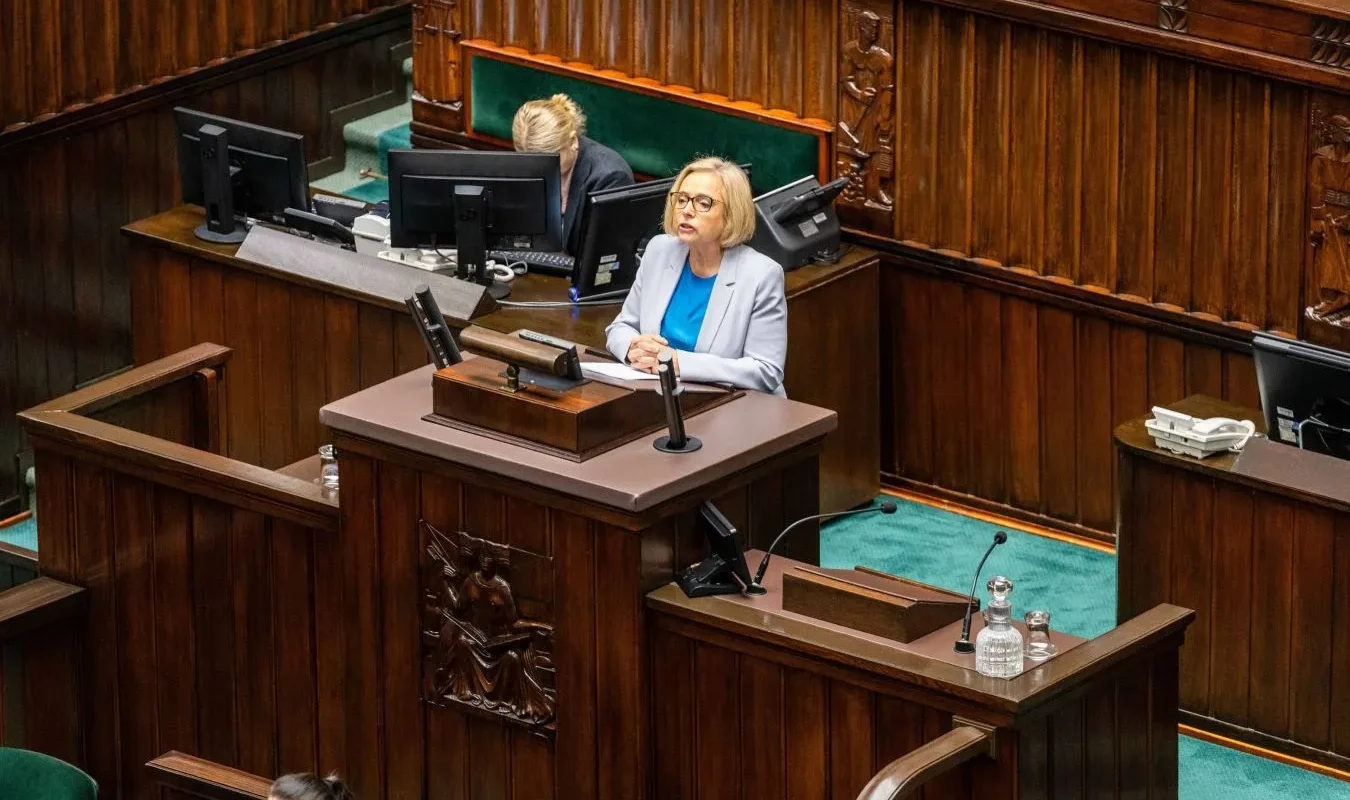 Posłanka wygłasza przemówienie z mównicy w Sejmie, w otoczeniu drewnianej architektury sali plenarnej i monitorów.