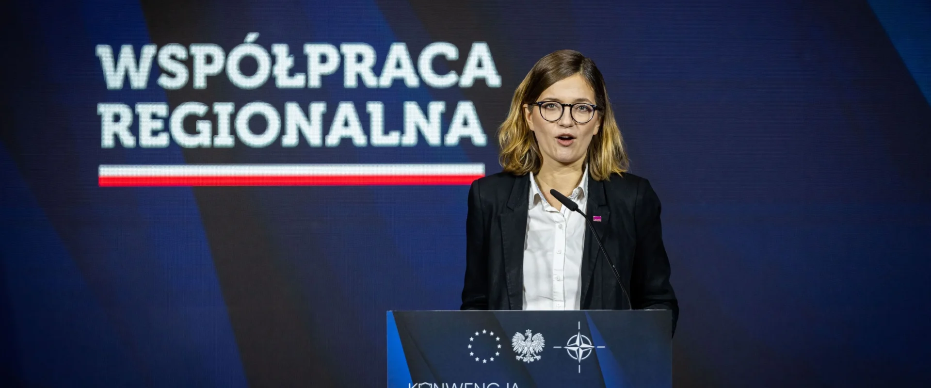 Kobieta z okularami stoi przy mównicy i wygłasza przemówienie podczas konferencji, na tle ekranu z napisem „WSPÓŁPRACA REGIONALNA” oraz banera „Konwencja Bezpieczeństwo i Współpraca”.