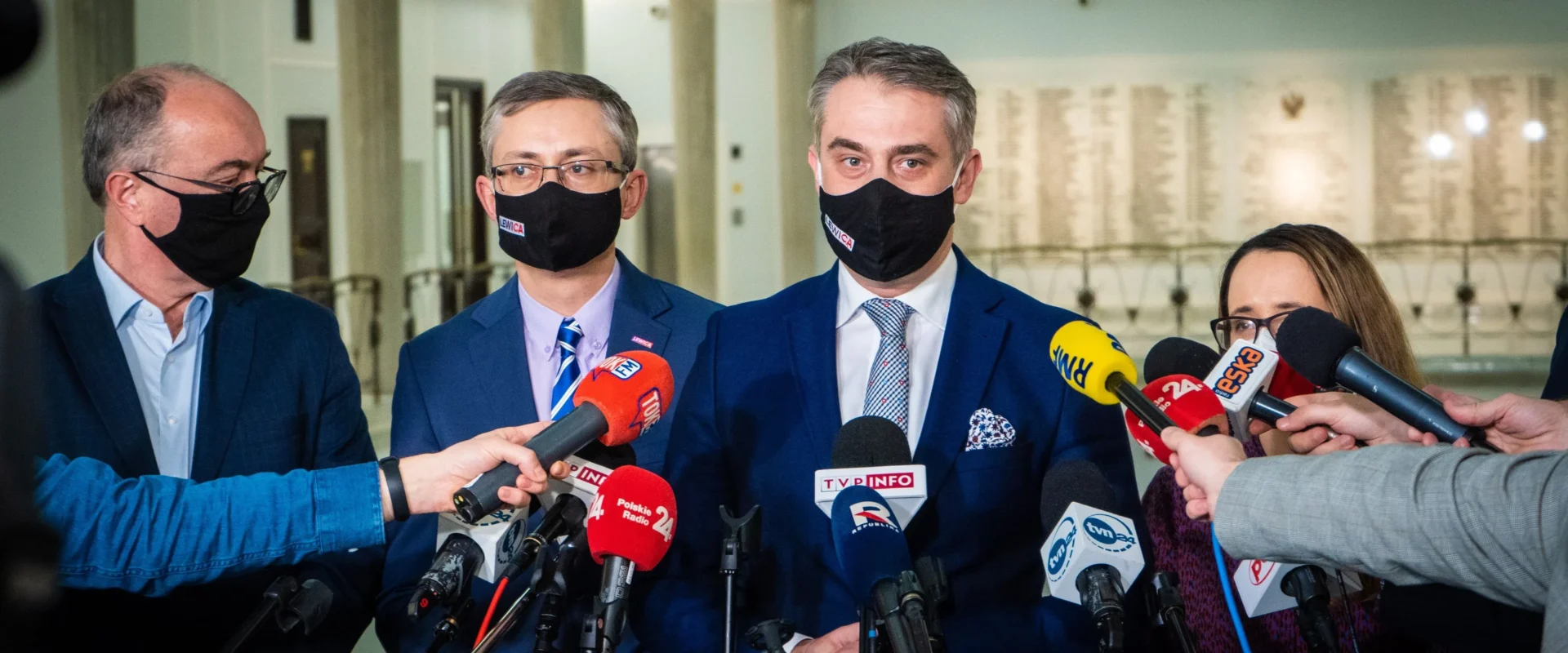 Na zdjęciu widać grupę mężczyzn i kobiet w garniturach, którzy udzielają wywiadu dziennikarzom biorącym udział w konferencji prasowej.