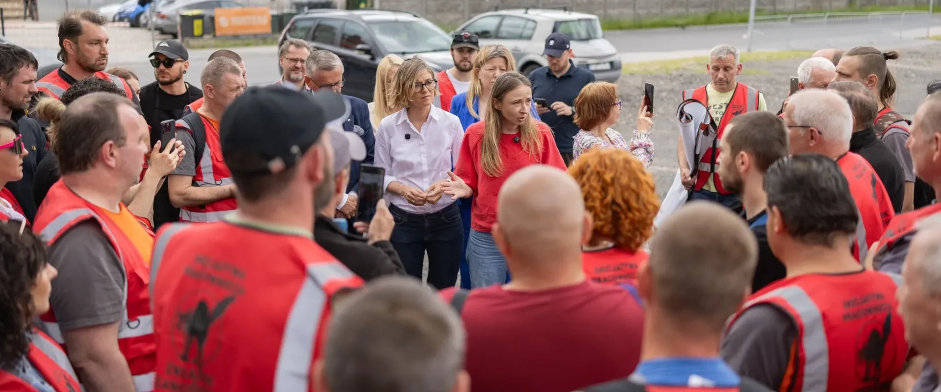 Grupa pracowników w czerwonych kamizelkach odblaskowych zgromadzona wokół dwóch kobiet przemawiających do tłumu podczas strajku w Jeremiasie.