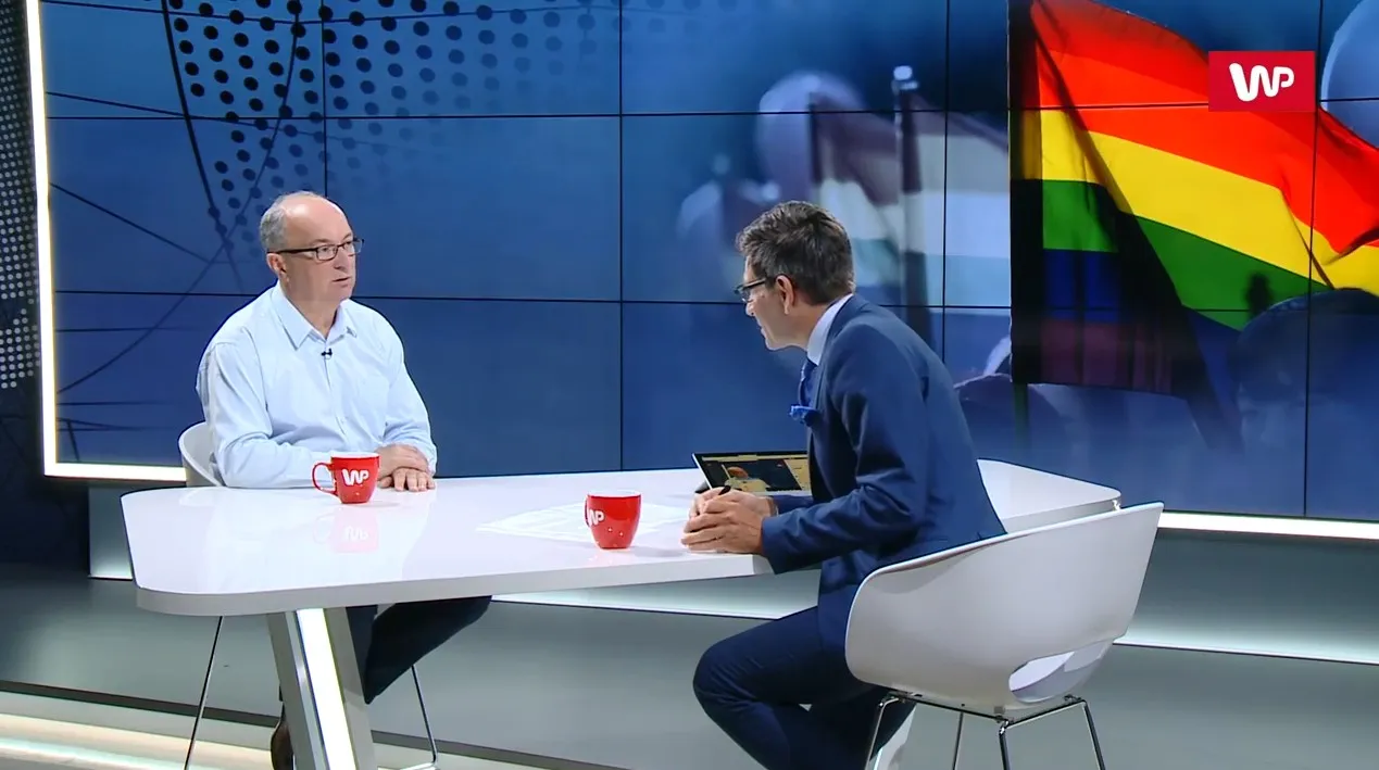 Dwóch mężczyzn rozmawia w studiu telewizyjnym przy białym stole; w tle ekran z tęczową flagą, a na stole czerwone kubki z logo WP.