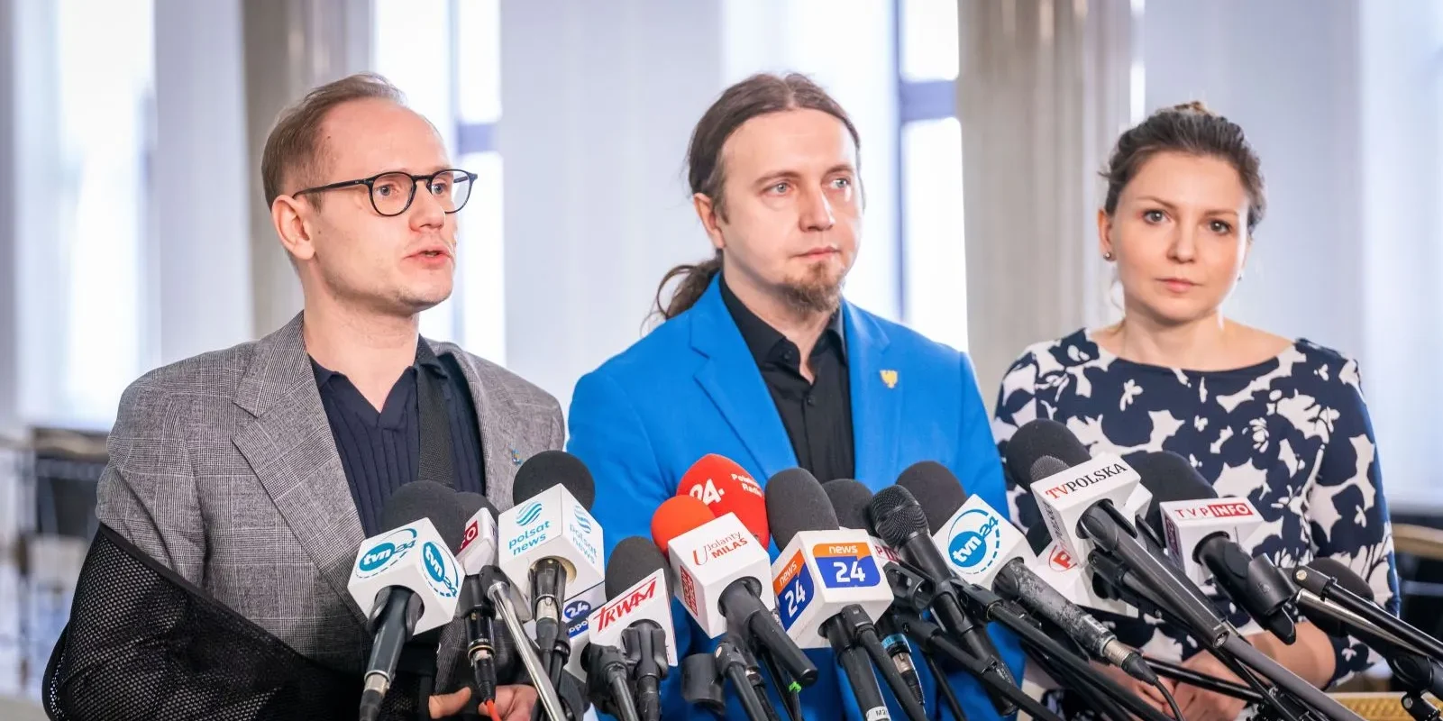 Trzy osoby stojące przy konferencji prasowej za licznymi mikrofonami: po lewej mężczyzna w szarym, kratkowanym garniturze z czarną koszulą, w środku mężczyzna w niebieskim żakiecie, a po prawej kobieta w granatowej sukience z białym wzorem.