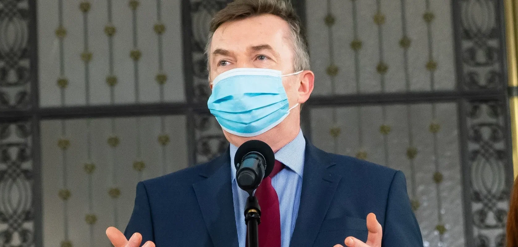 Mężczyzna w garniturze, jasnoniebieskiej koszuli i czerwonym krawacie z niebieską maseczką ochronną na twarzy przemawia do mikrofonu.