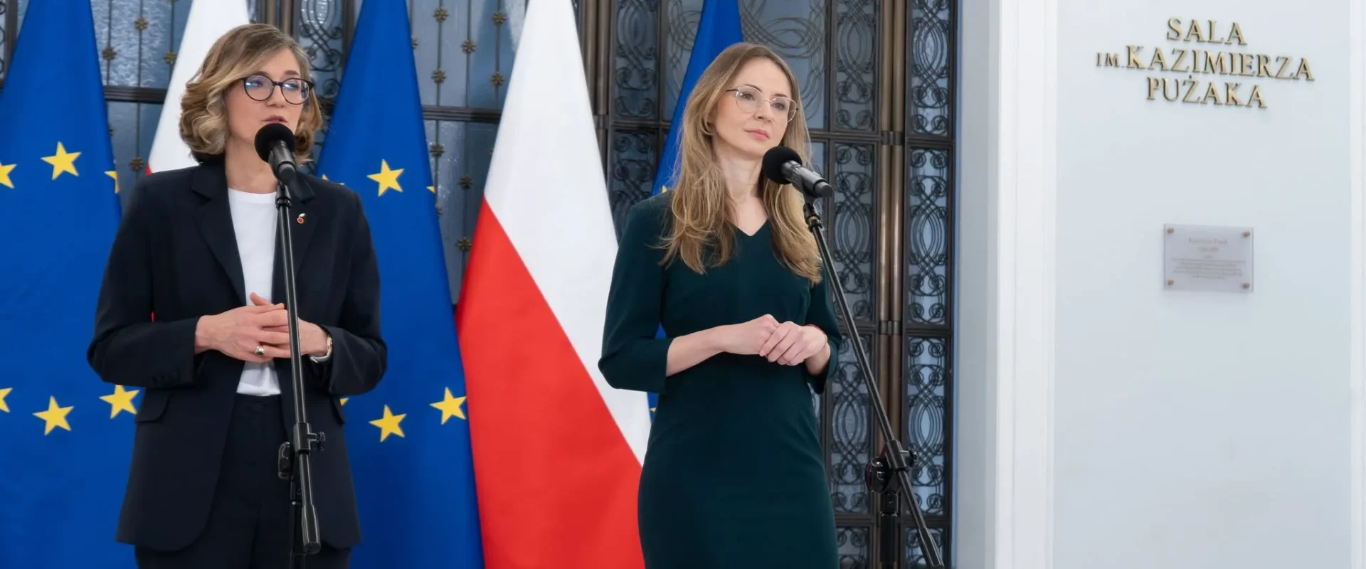Dwie kobiety przemawiające do mikrofonów podczas konferencji prasowej przed wejściem do Sali im. Kazimierza Pużaka, otoczone flagami Unii Europejskiej i Polski, w kontekście postulatu likwidacji Funduszu Kościelnego.