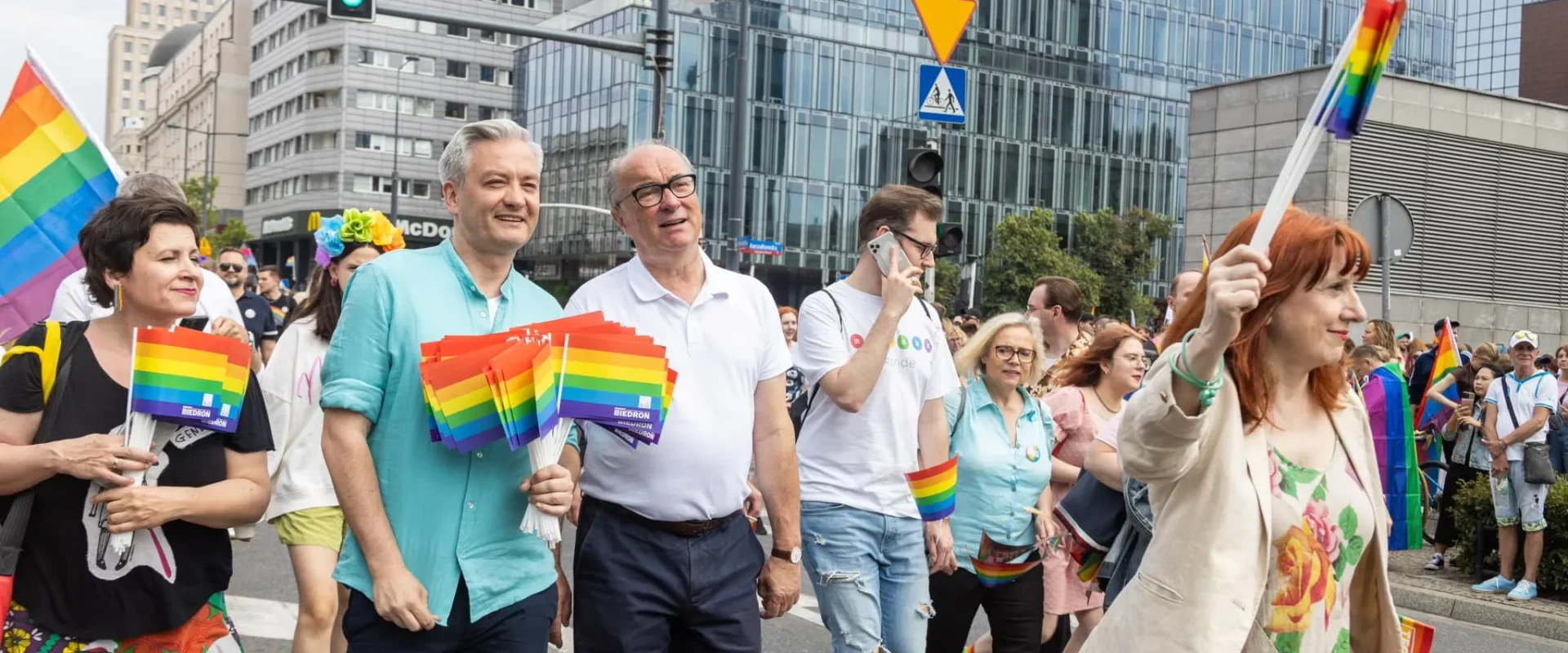 Uczestnicy Parady Równości 2023 maszerują ulicą miasta, trzymając tęczowe flagi i plakietki, wśród wysokich nowoczesnych budynków.