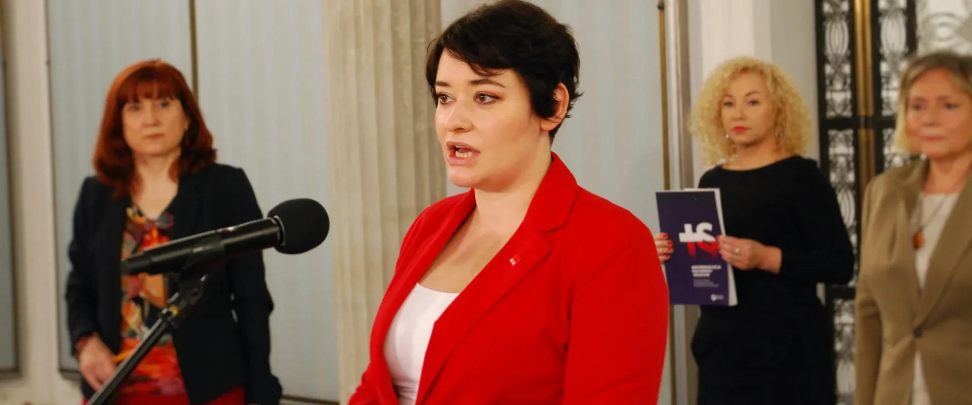 Kobieta w czerwonym żakiecie przemawia do mikrofonu podczas konferencji, w tle stoją trzy inne kobiety.