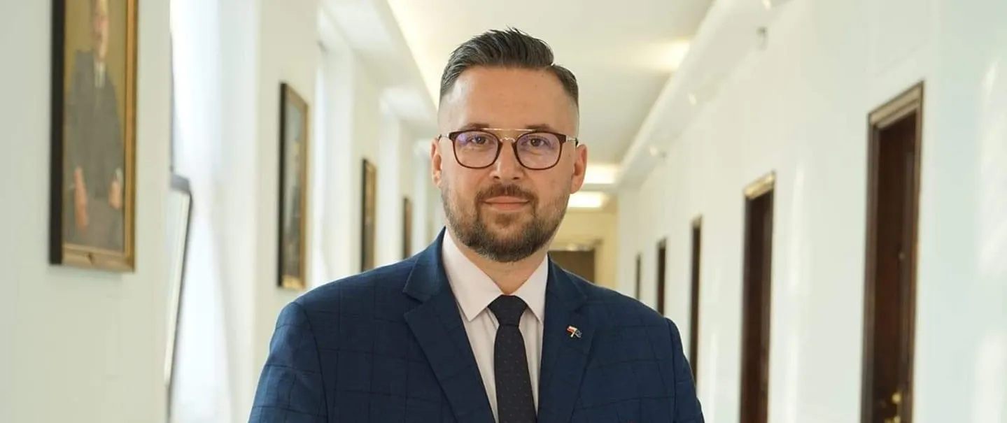 Mężczyzna w granatowym garniturze i okularach stoi w jasnym, długim korytarzu biurowym, patrząc w kamerę.