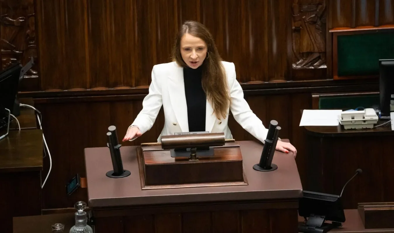 Kobieta w białej marynarce przemawia z mównicy w parlamencie, wyrażając swoje zdanie na ważny temat.