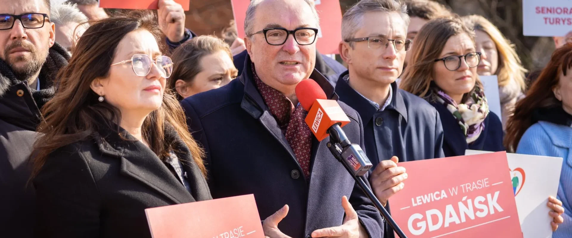 Na zdjęciu widoczna jest grupa osób podczas konferencji prasowej, w której uczestniczy mężczyzna z mikrofonem oraz kobiety i mężczyźni trzymający transparenty z napisem "LEWICA W TRASIE GDAŃSK".