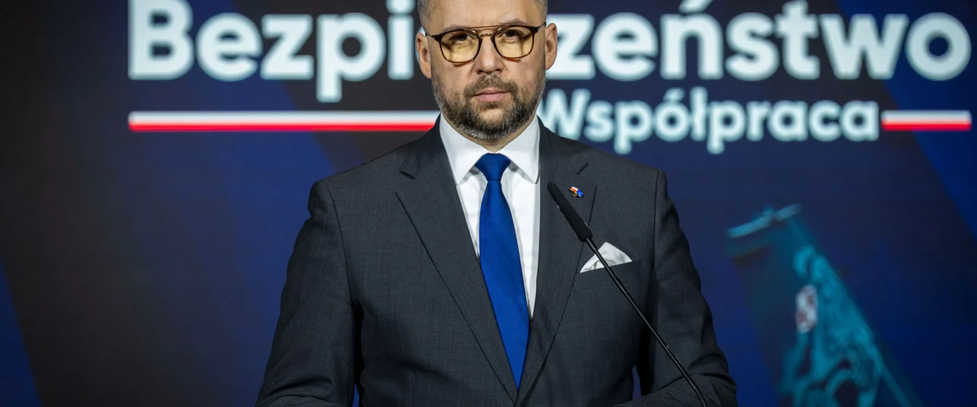 Mężczyzna w garniturze i okularach stoi za mównicą podczas konferencji o bezpieczeństwie i współpracy, na tle widoczne są napisy „Bezpieczeństwo” i „Współpraca”.