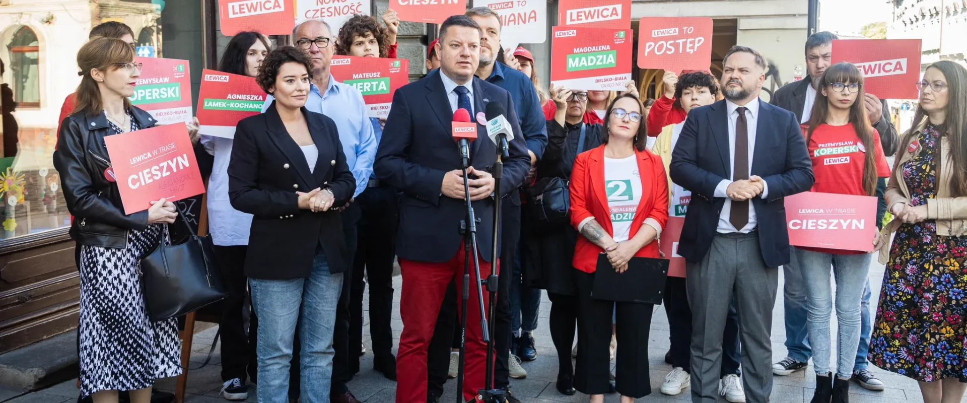 Grupa osób na ulicznym wiecu politycznym; w centrum mężczyzna w garniturze przemawia do mikrofonów, otoczony innymi uczestnikami trzymającymi czerwone plakaty Lewica.