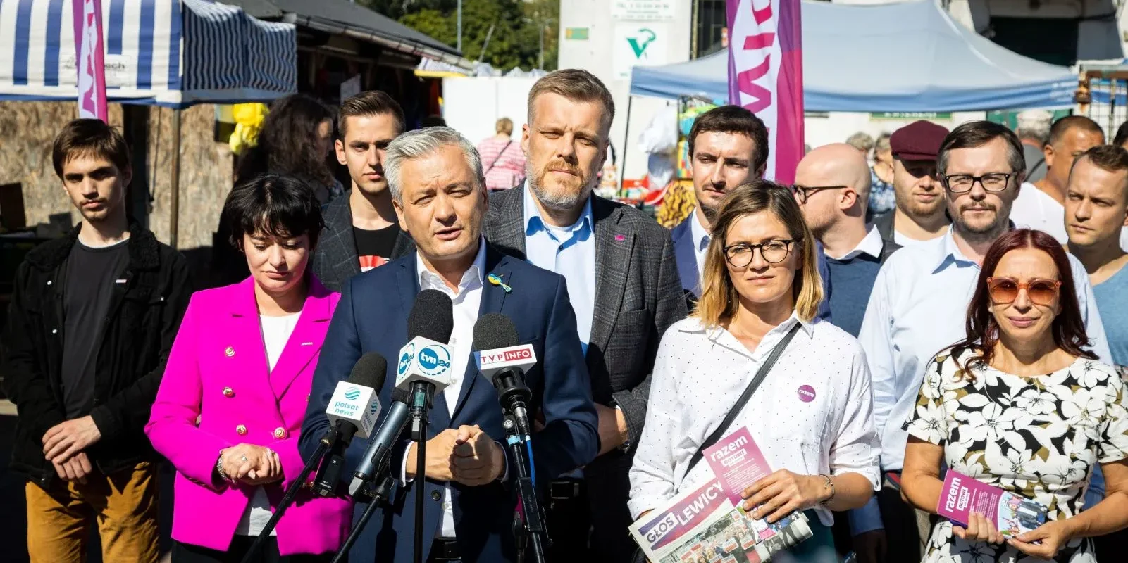 Grupa osób na konferencji prasowej na targowisku; mężczyzna w granatowym garniturze przemawia do mikrofonów, otoczony przez reporterów i innych uczestników.