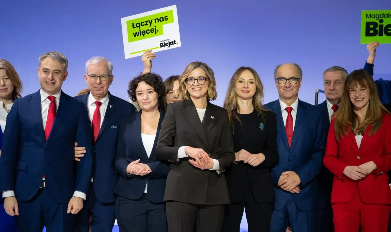Grupa osób w eleganckich garniturach i marynarkach stoi na scenie na tle niebieskiego tła, trzymając plakat z hasłem „Łączy nas więcej”.