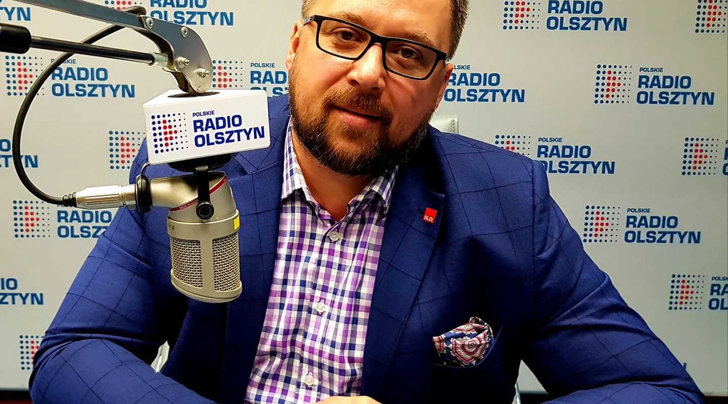 Mężczyzna z brodą i okularami, ubrany w granatowy garnitur i koszulę w kratę, siedzi przy mikrofonie w studiu radiowym z logami Polskiego Radia Olsztyn w tle.