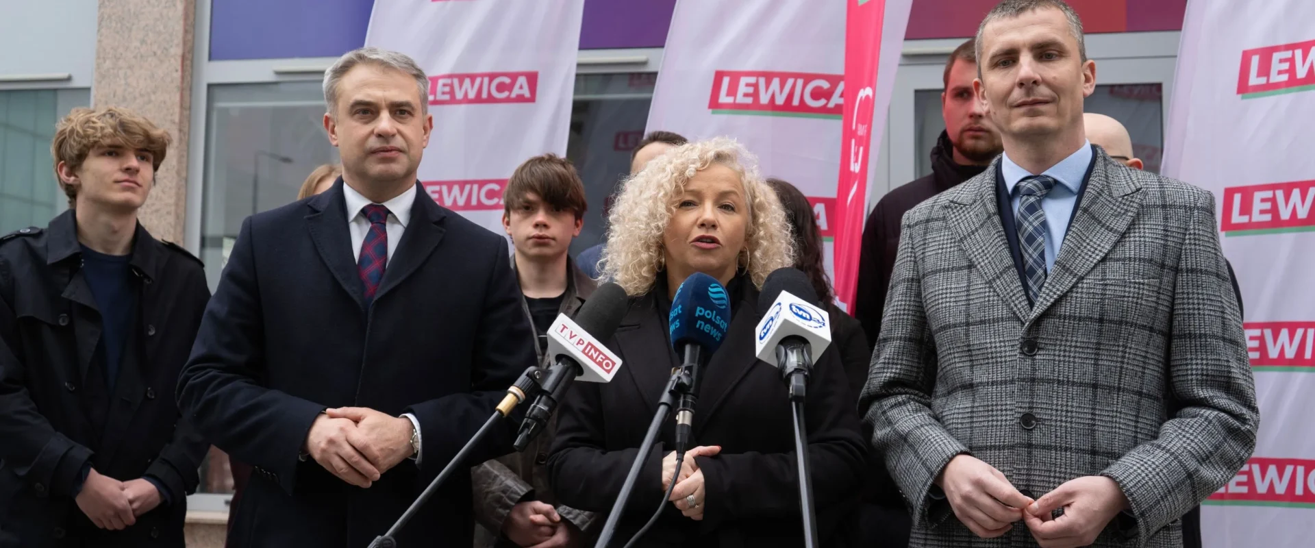 Grupa osób podczas konferencji prasowej Lewicy, stojących przed mikrofonami na tle flag z napisem LEWICA.