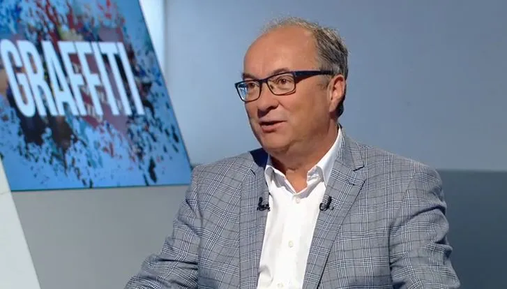 Mężczyzna w okularach, w garniturze w kratę i białej koszuli, rozmawia w studiu telewizyjnym; w tle widać ekran z napisem „GRAFFITI”.