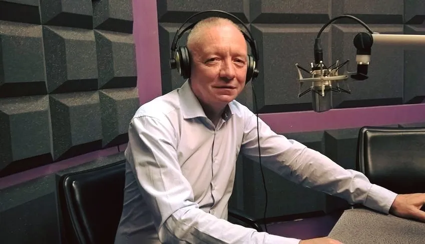 Mężczyzna w białej koszuli siedzi w studiu radiowym przy stole z mikrofonem i słuchawkami.