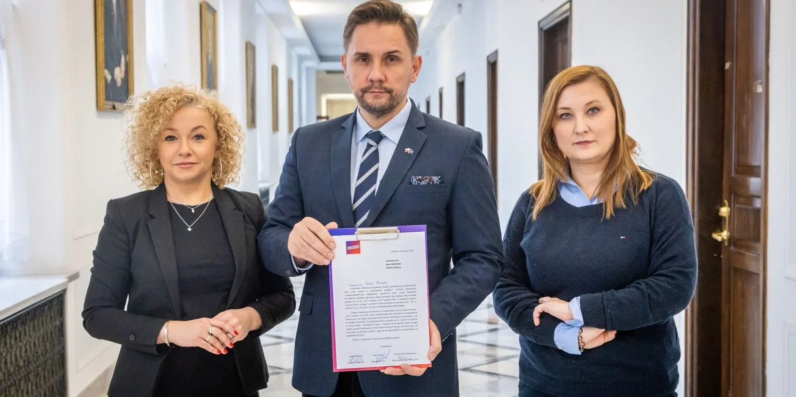 Trzy osoby stoją w korytarzu, trzymając dokument, na tle eleganckiej architektury z obrazami na ścianach.