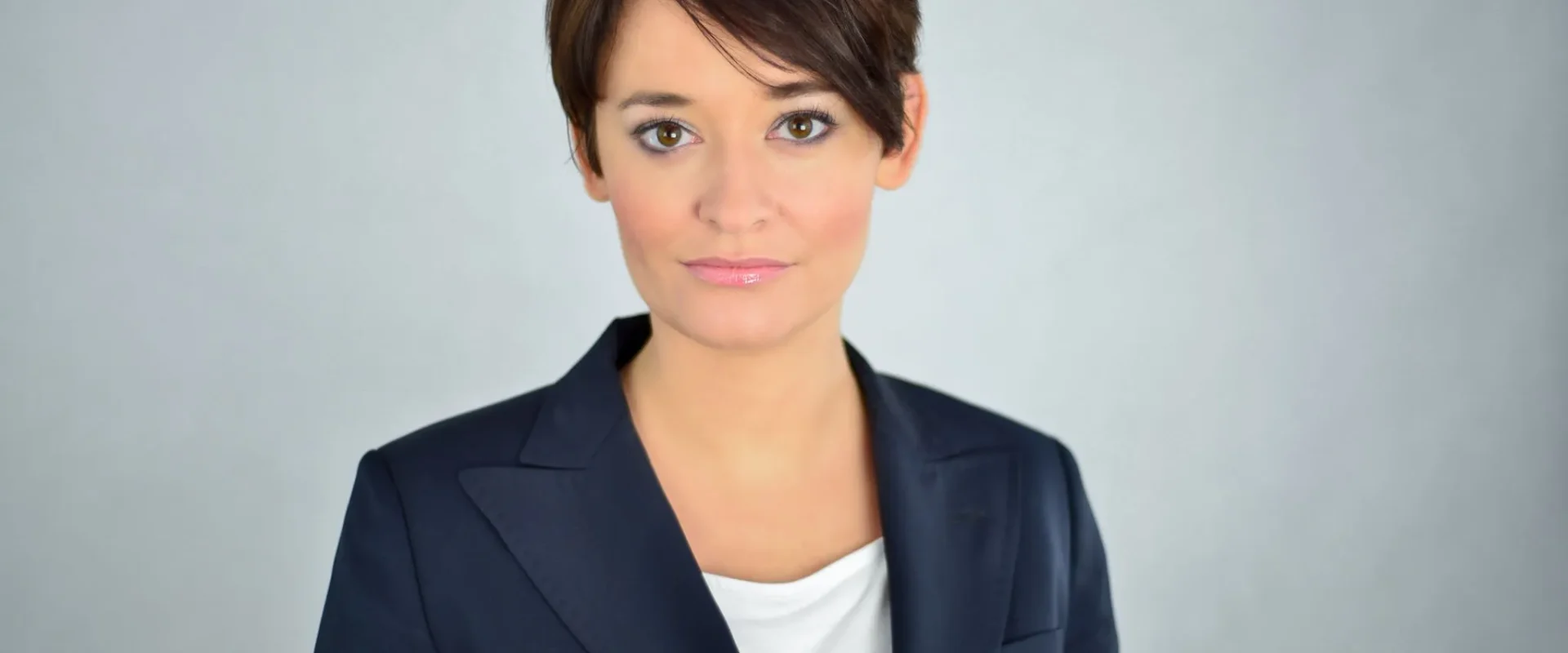 Portret kobiety z krótkimi brązowymi włosami, ubranej w granatowy żakiet i biały top, na neutralnym jasnym tle.