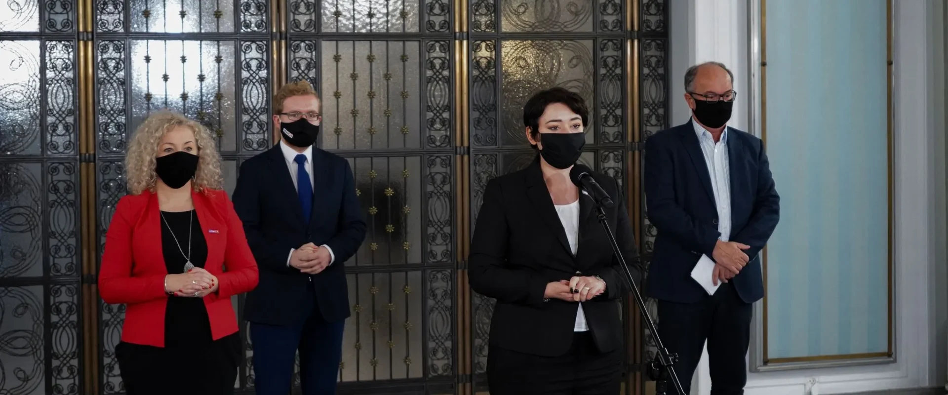 Kobieta w czarnym garniturze przemawia do mikrofonu podczas konferencji prasowej, stojąc między trzema osobami w garniturach, wszyscy w maseczkach, na tle ozdobnej metalowej bramy.