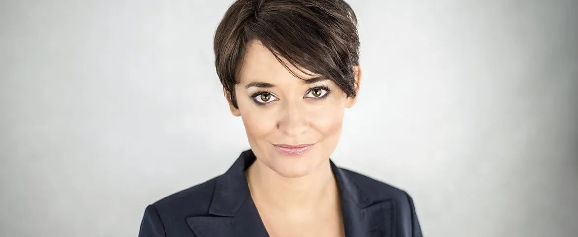 Portret uśmiechniętej kobiety o krótkich ciemnych włosach, w granatowej marynarce na jasnym, rozmytym tle.
