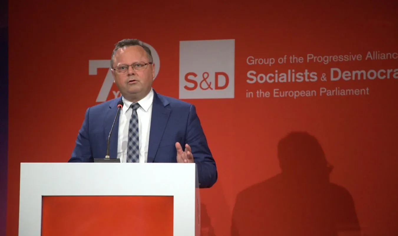 Mężczyzna w granatowym garniturze przemawia z mównicy na czerwonym tle z logiem S&D (Socialists & Democrats) w Parlamencie Europejskim.