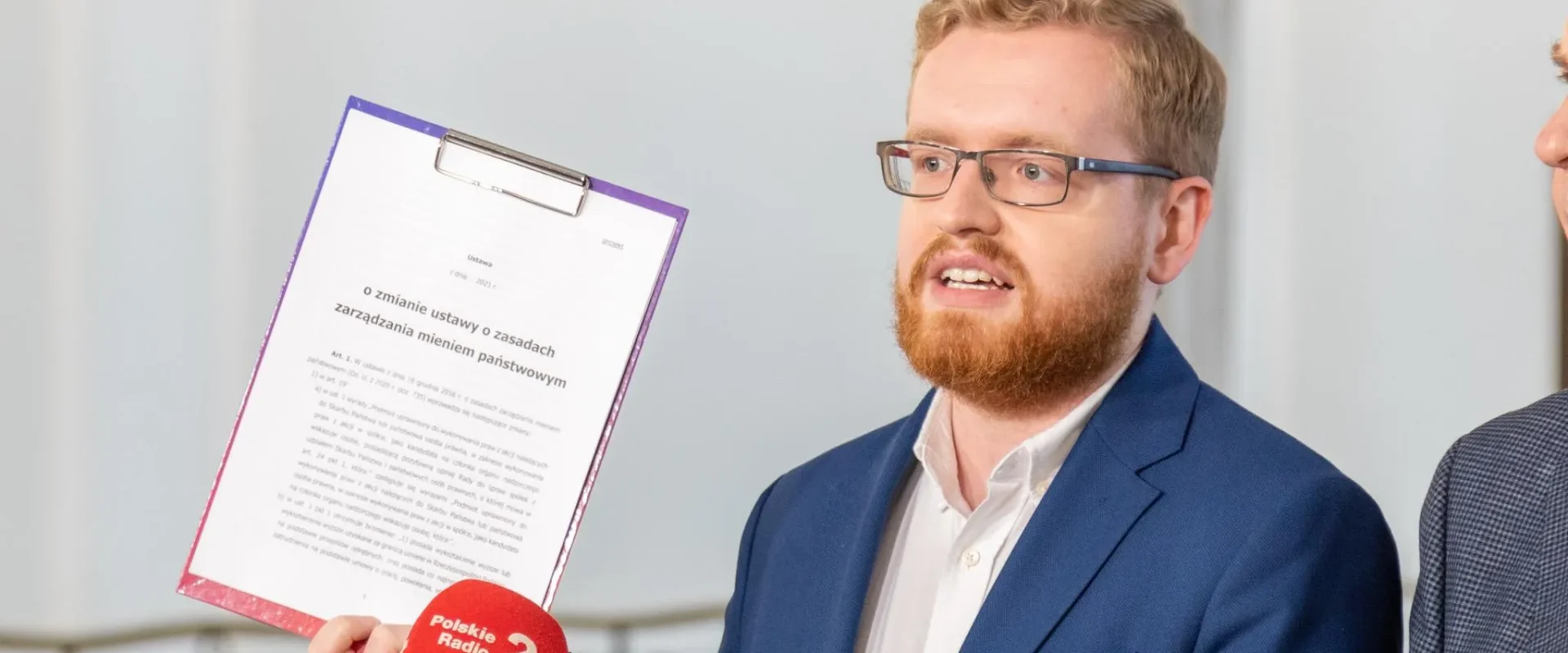 Mężczyzna z brodą i okularami w granatowym garniturze prezentuje projekt podatku cyfrowego podczas konferencji prasowej, trzymając dokument na klipboardzie i otoczony mikrofonami, projekt ma być gotowy w styczniu.