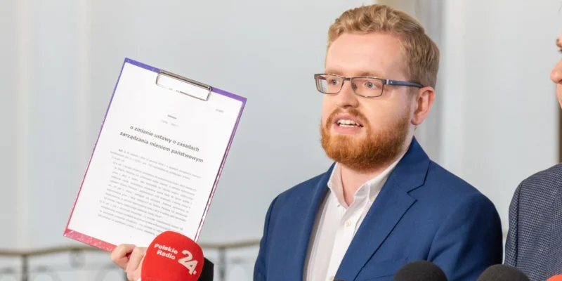 Mężczyzna z brodą i okularami w granatowym garniturze prezentuje projekt podatku cyfrowego podczas konferencji prasowej, trzymając dokument na klipboardzie i otoczony mikrofonami, projekt ma być gotowy w styczniu.
