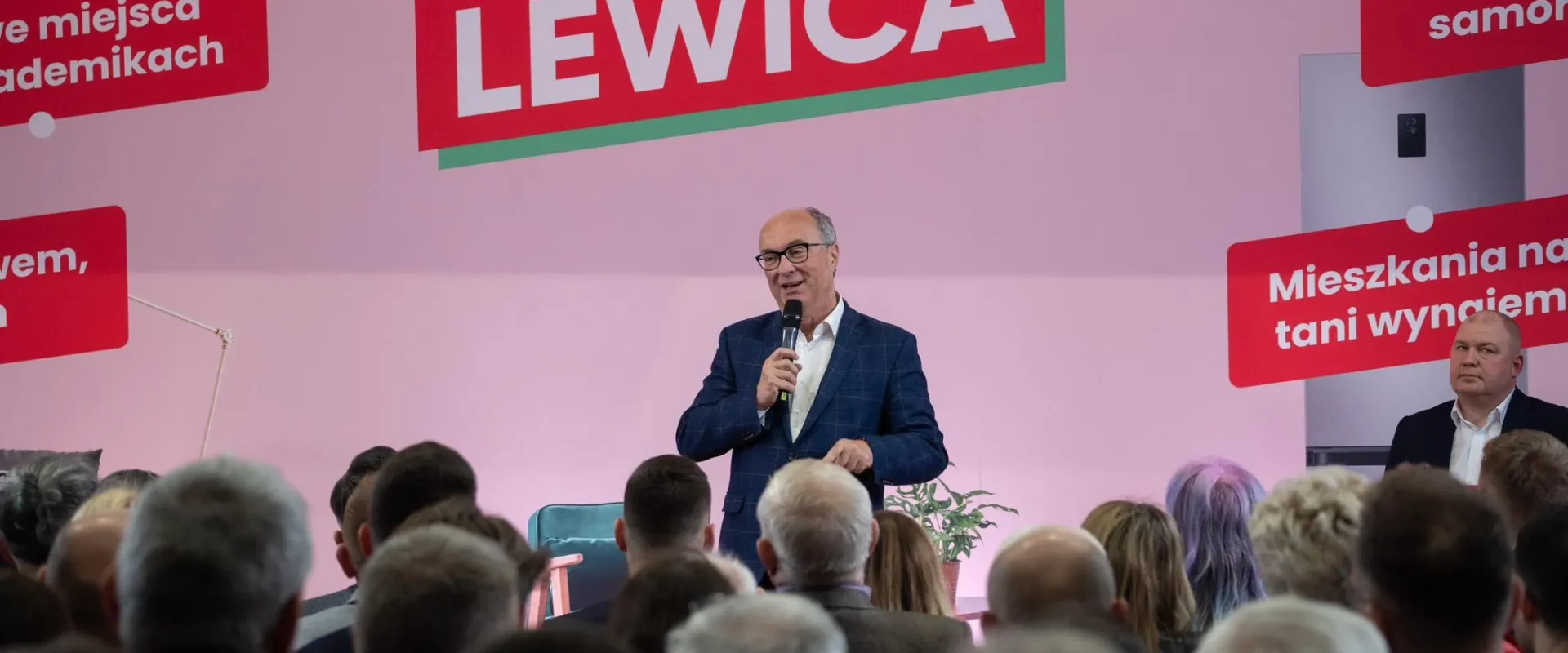 Mężczyzna w granatowym garniturze przemawia do zgromadzonej publiczności na scenie, na różowym tle z dużym napisem LEWICA.